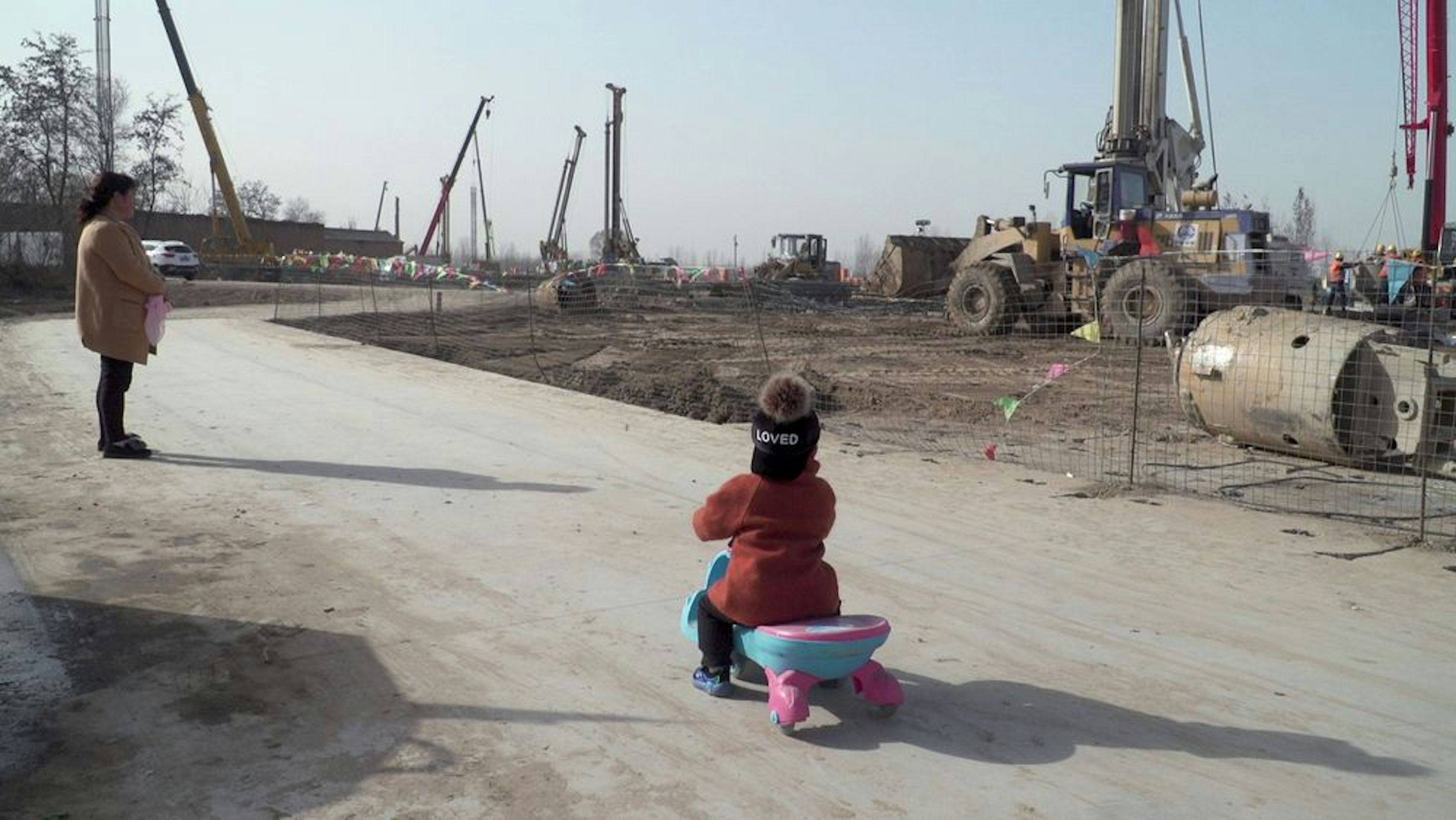 A woman and child stand and watch a construction site. The child is on a bike and wears a hat that says “Loved”.