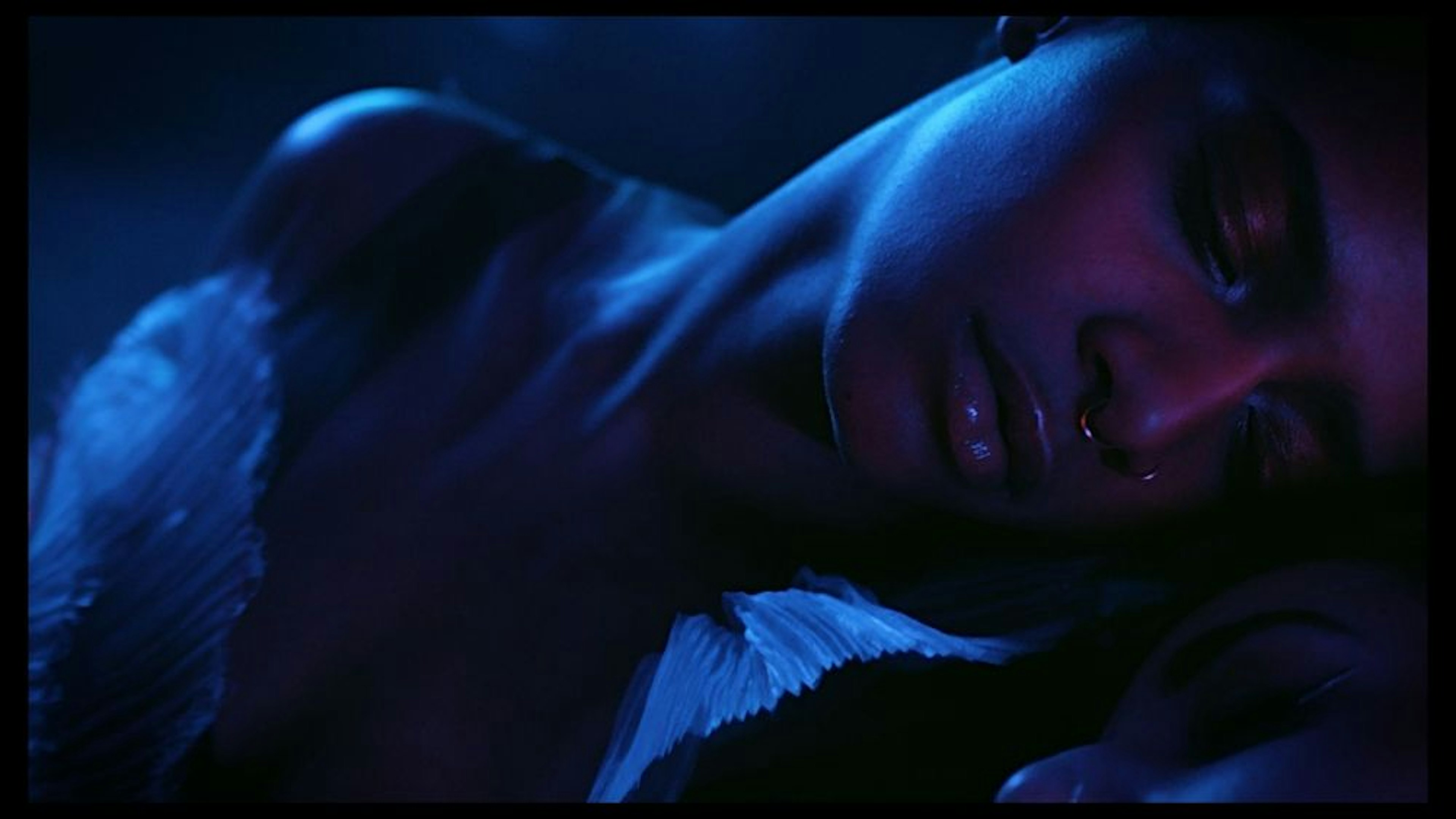 Close-up of a dark skinned woman resting her head gently on another person who is slightly visible. They are both lit by soft red and blue tones with a white key light illuminating the sides of their faces. 