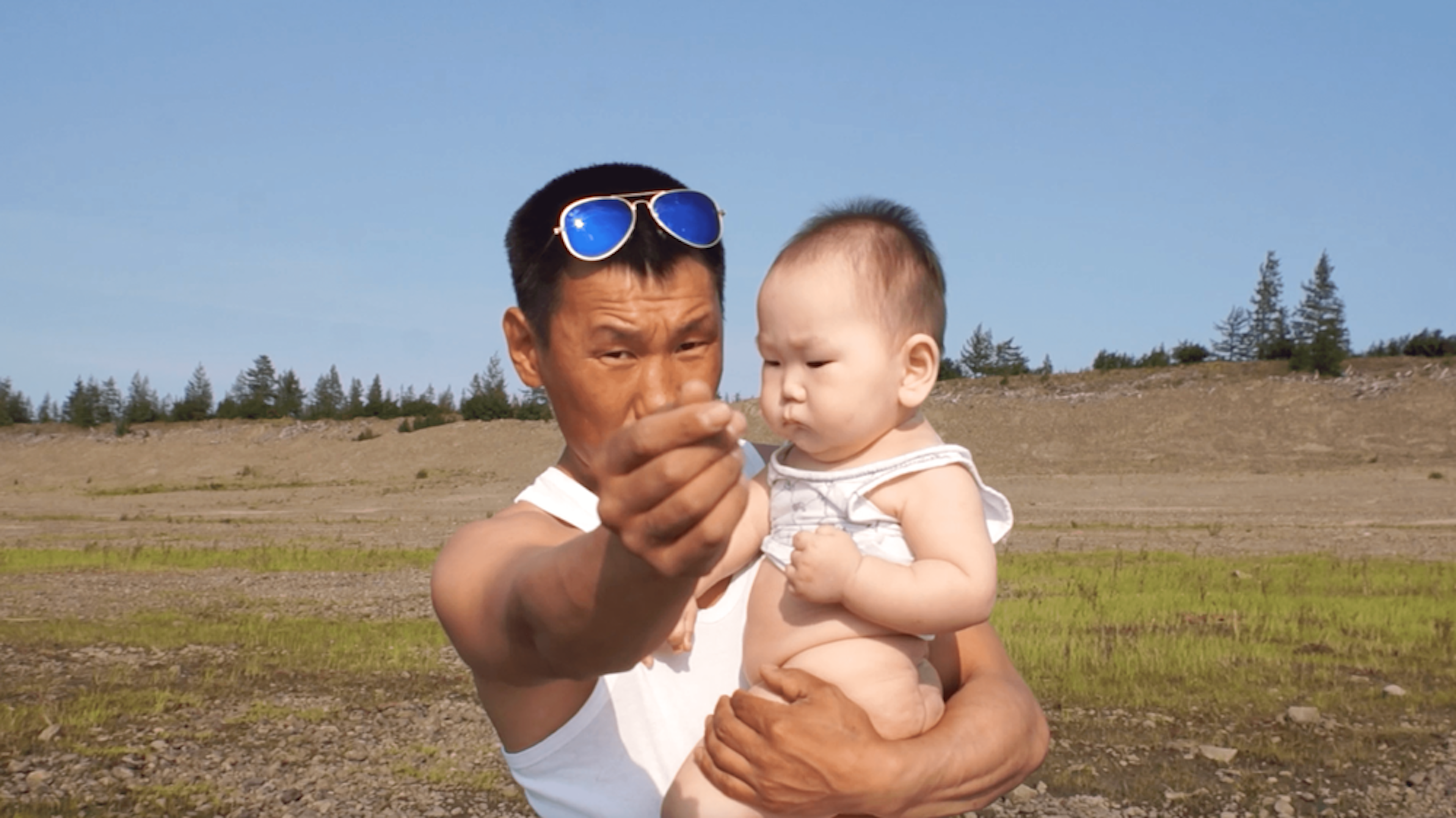 Man wearing blue-tinted sunglasses on his head holds a baby, his free hand close to the lens with thumb and index finger appearing to rub against each other. They are outside in a field on a blue, sunny day.