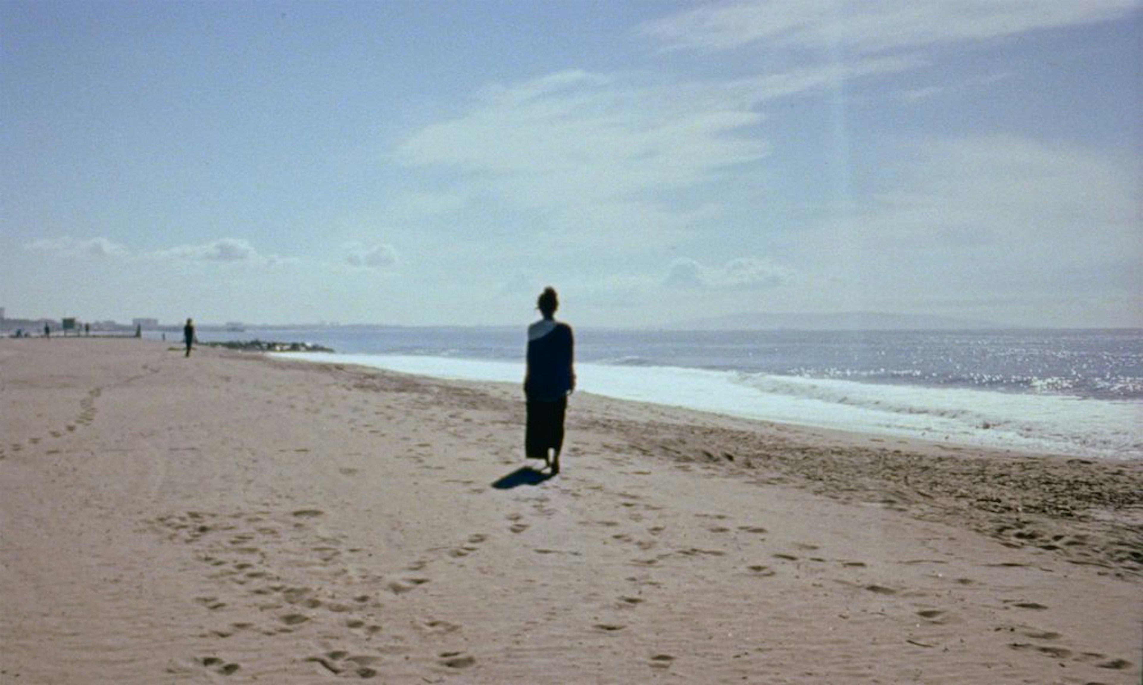 Landscape of a shoreline on a blue sunny day, footsteps visible in the sand and a figure whose back is turned toward the viewer. 