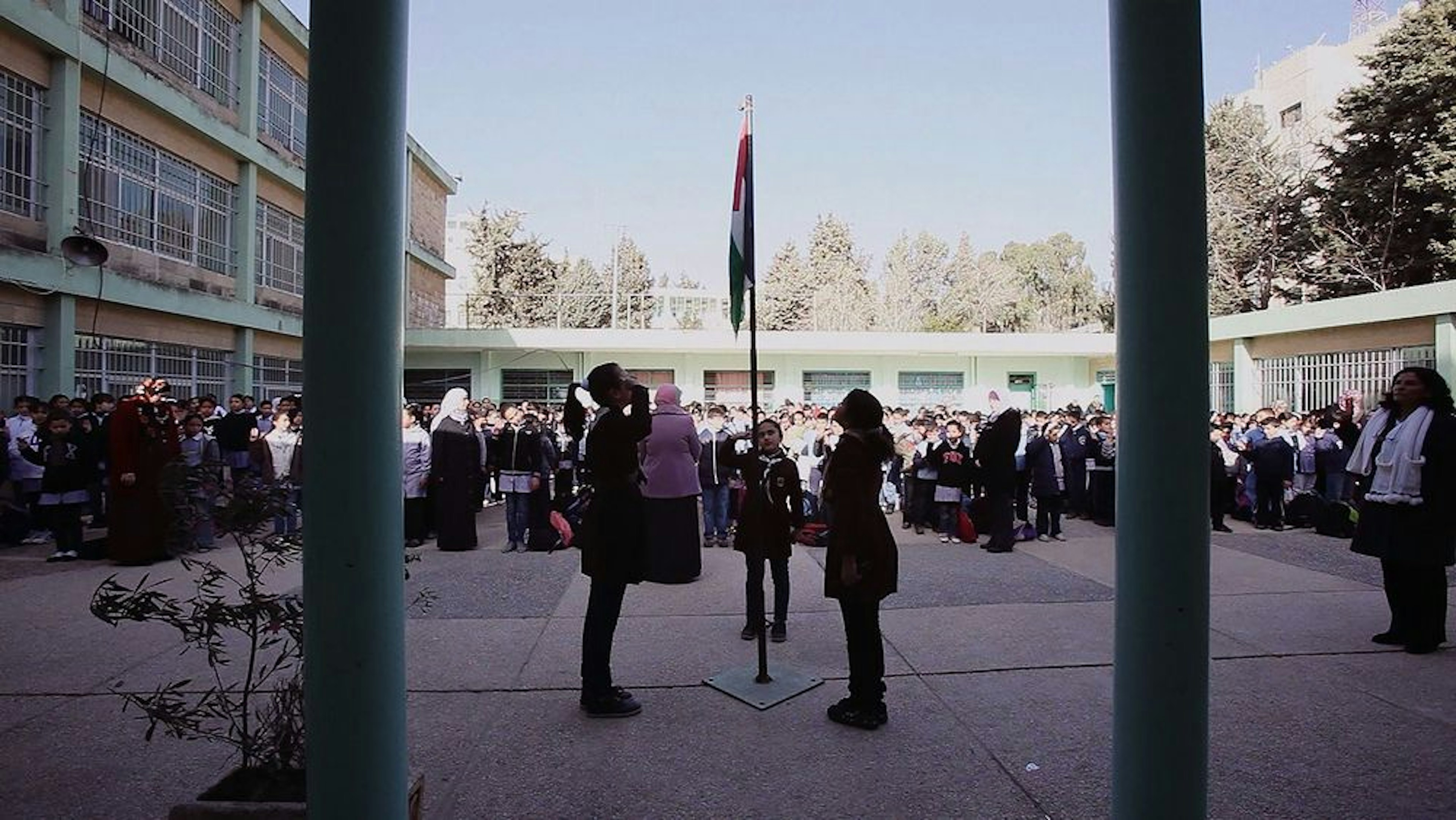 Outdoor courtyard with a crowd of people witnessing a trio of people featuring a salute to the Palestinian flag—green, red and white—set atop a flagpole. 
