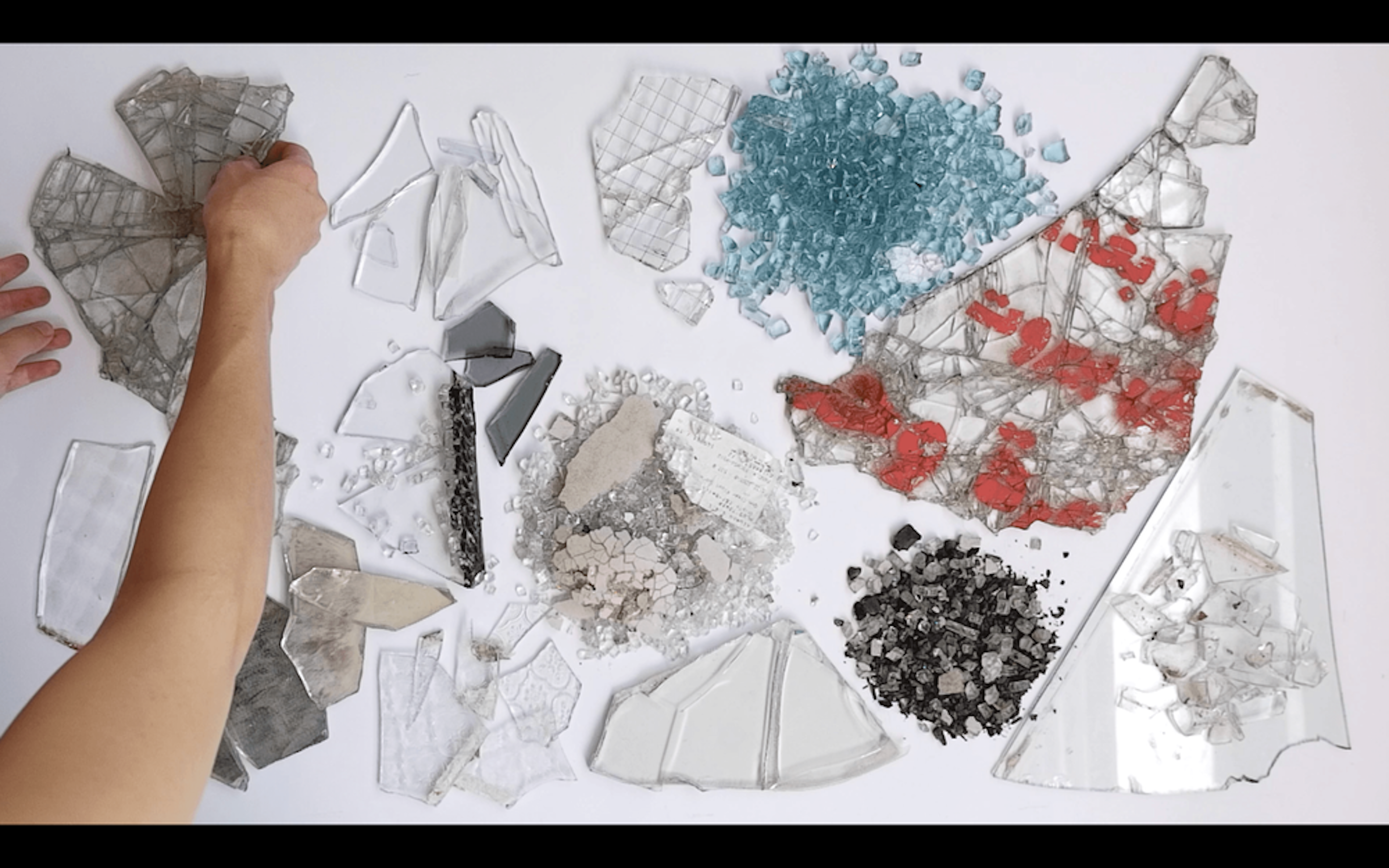 Overhead view of various pieces of colorful broken glass, assembled neatly. There is an arm on the left side arranging pieces of glass.