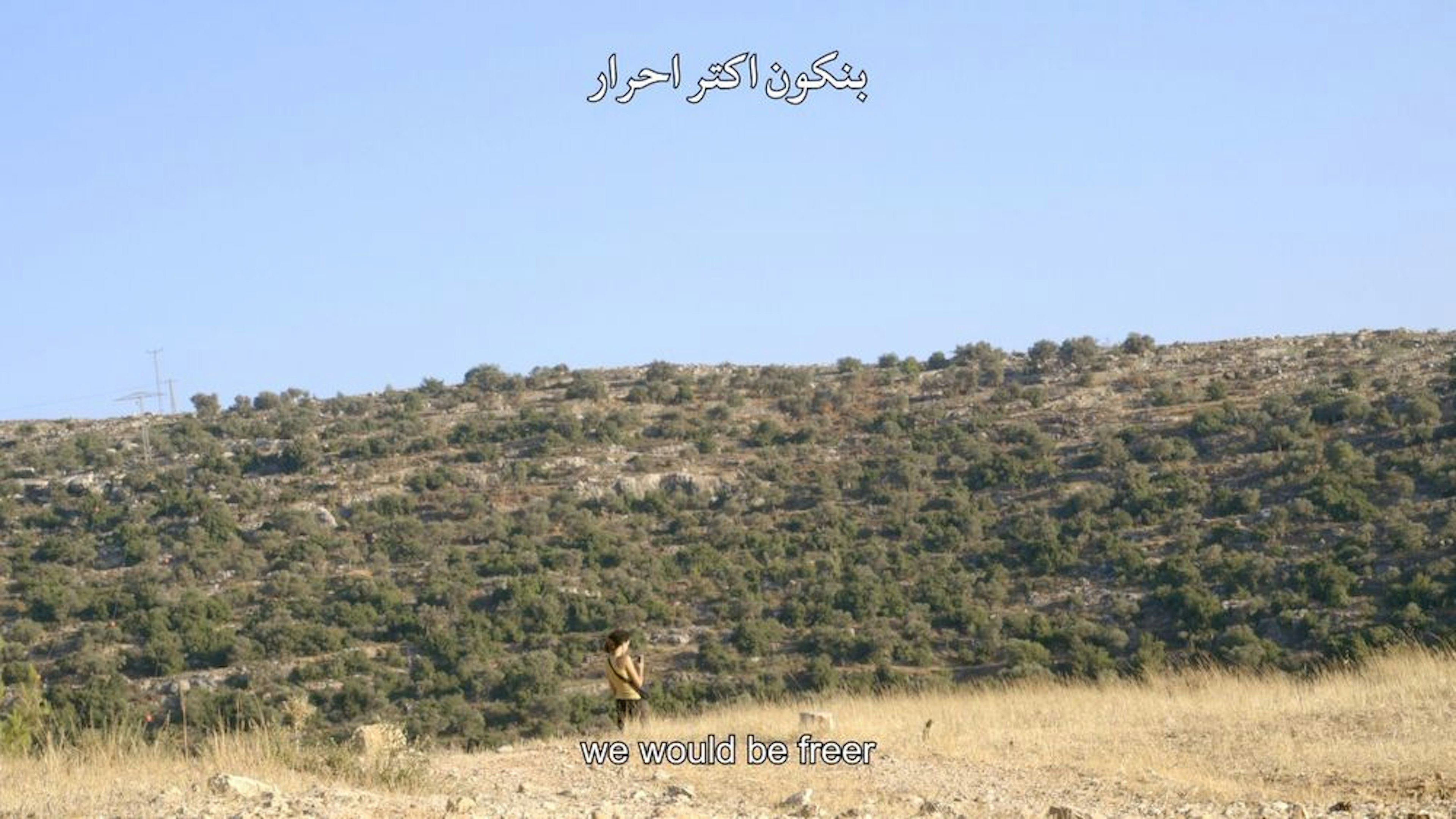 Blue sky and sunny day illuminating a landscape on the side of a hill. A small figure is seen in the distance. On the top is writing in Arabic letters. On bottom, subtitles read, “We would be freer.”
