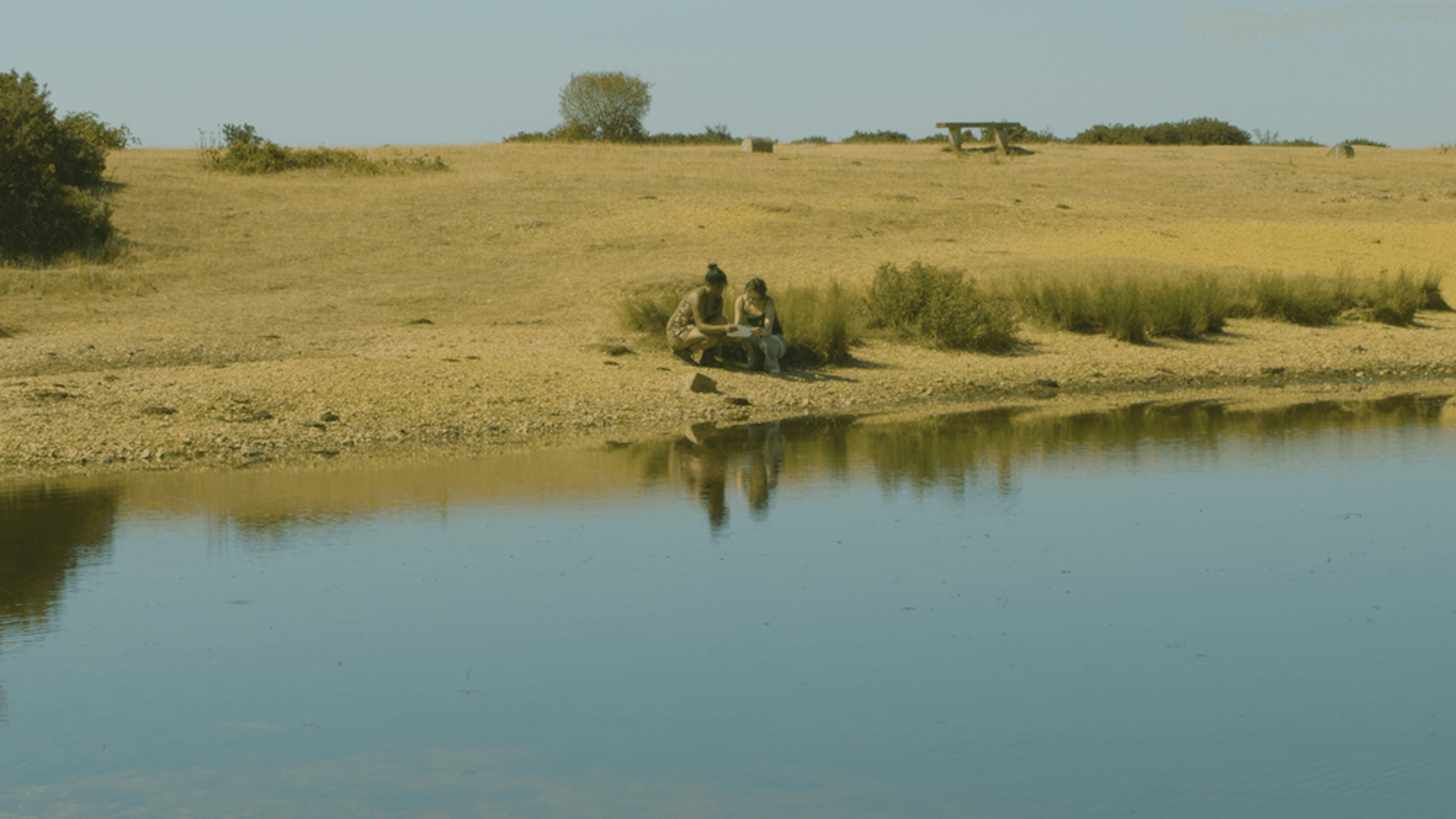 a wide shot of a landscape. two persons are sitting by the water looking at a piece of paper 