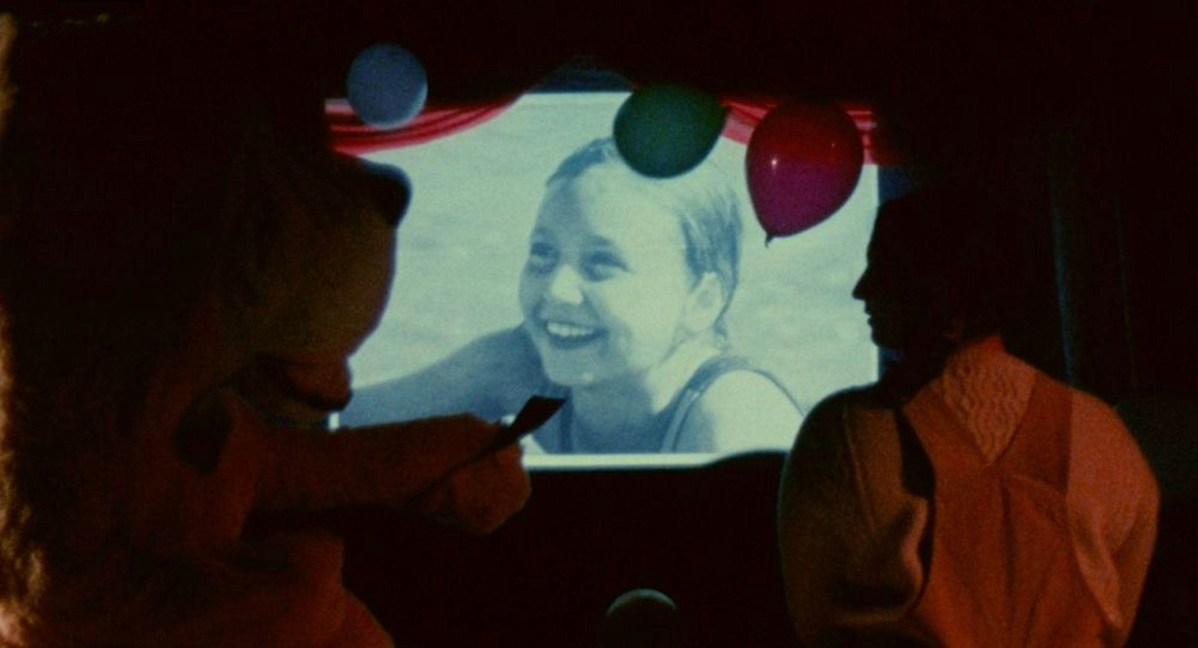 Films still showing a person and a teddy bear standing in front of a screen depicting a monochrome portrait of a child smiling, surrounded with balloons 