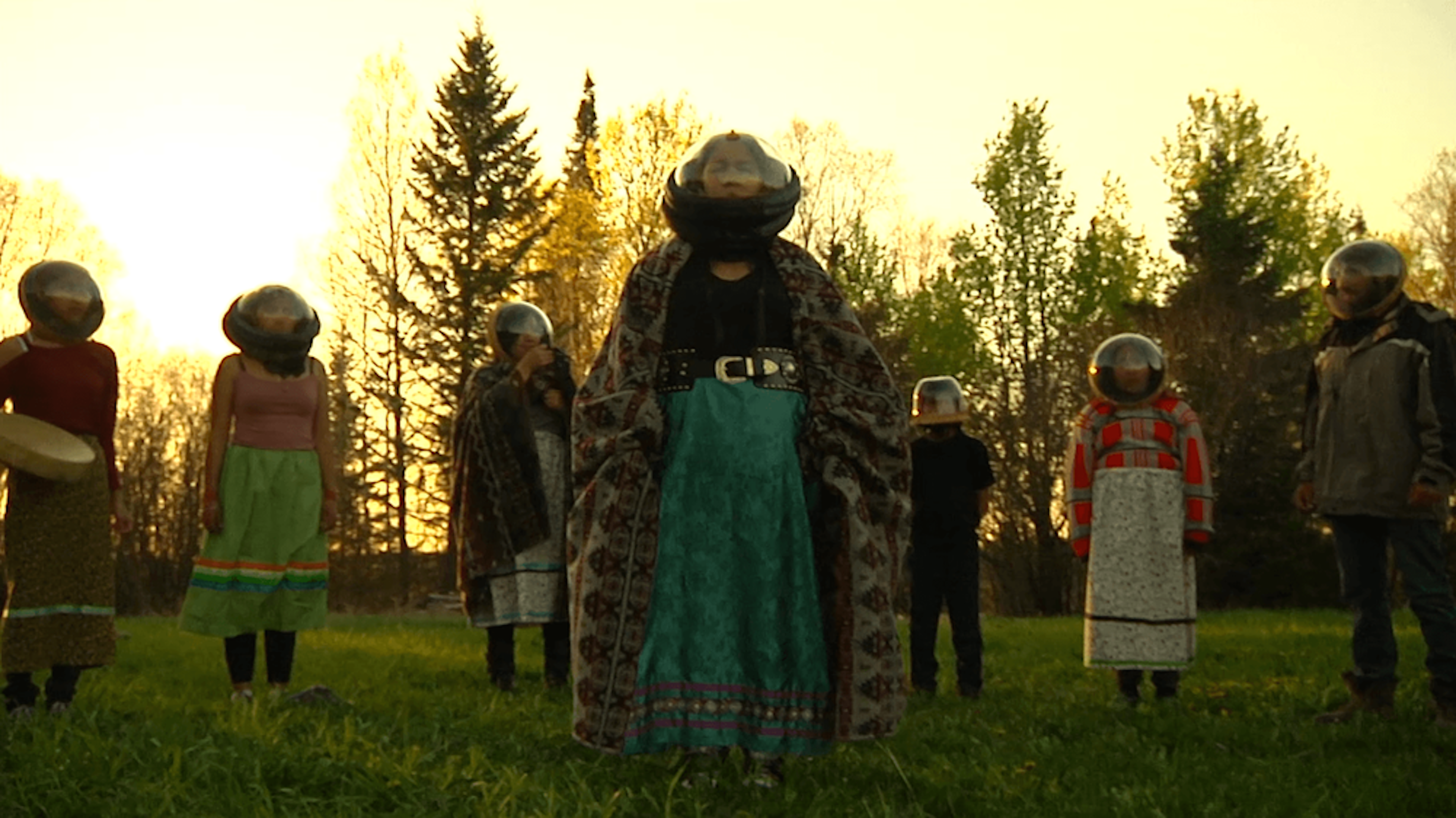 Six people stand in a semicircle with a seventh person in the middle. They are surrounded by trees and grass with the sun setting behind them.