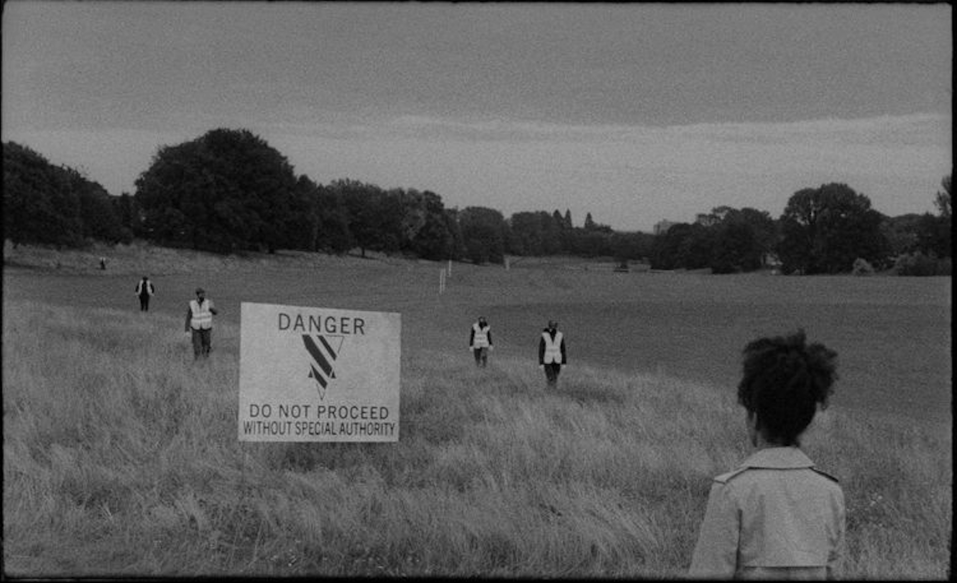 Black and white film still of a field, on the left a large sign that reads “Danger do not proceed without special authority”. There is a person in the foreground looking at a group of people wearing safety vests behind the sign.