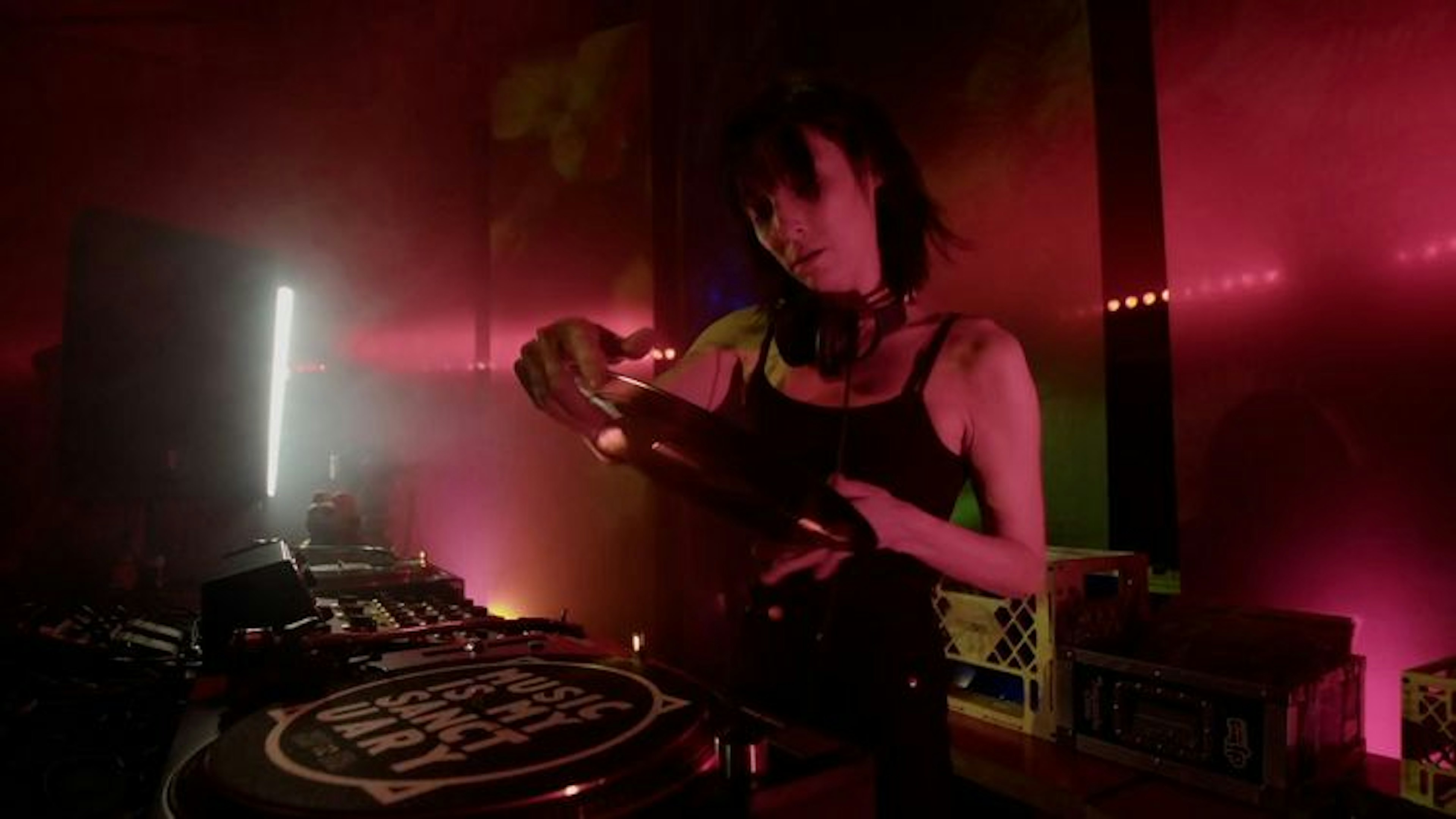 Diana in front of a DJ booth lit in dim red light holding a 12" record. She wears a black tank top and headphones around her neck.