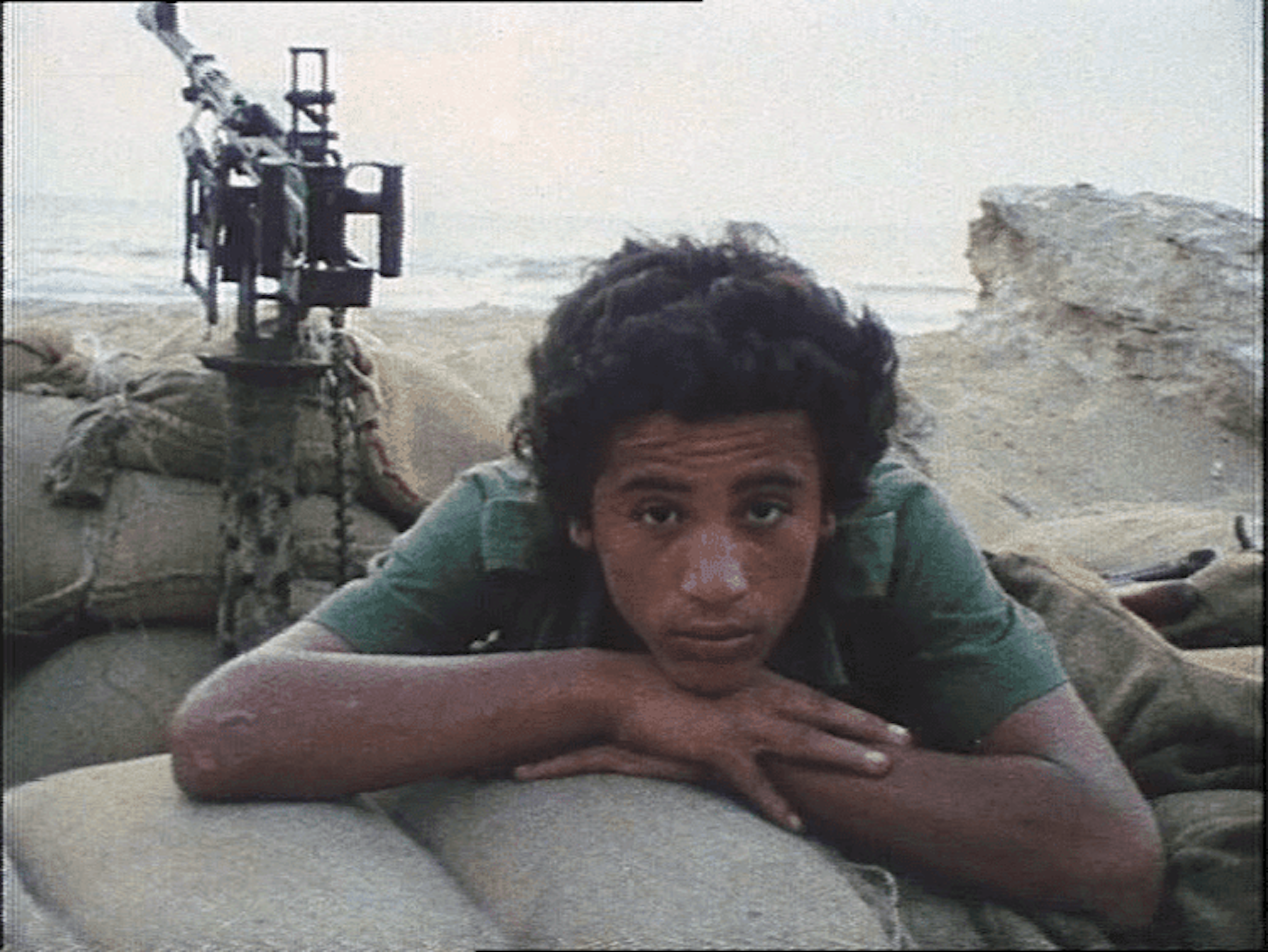 A dark-skinned man gazes at the camera, his chin resting on his hands resting atop sandbags. He wears a short sleeved green shirt. Behind him is military equipment facing the background. There is a shoreline in the background and a hazy sun.
