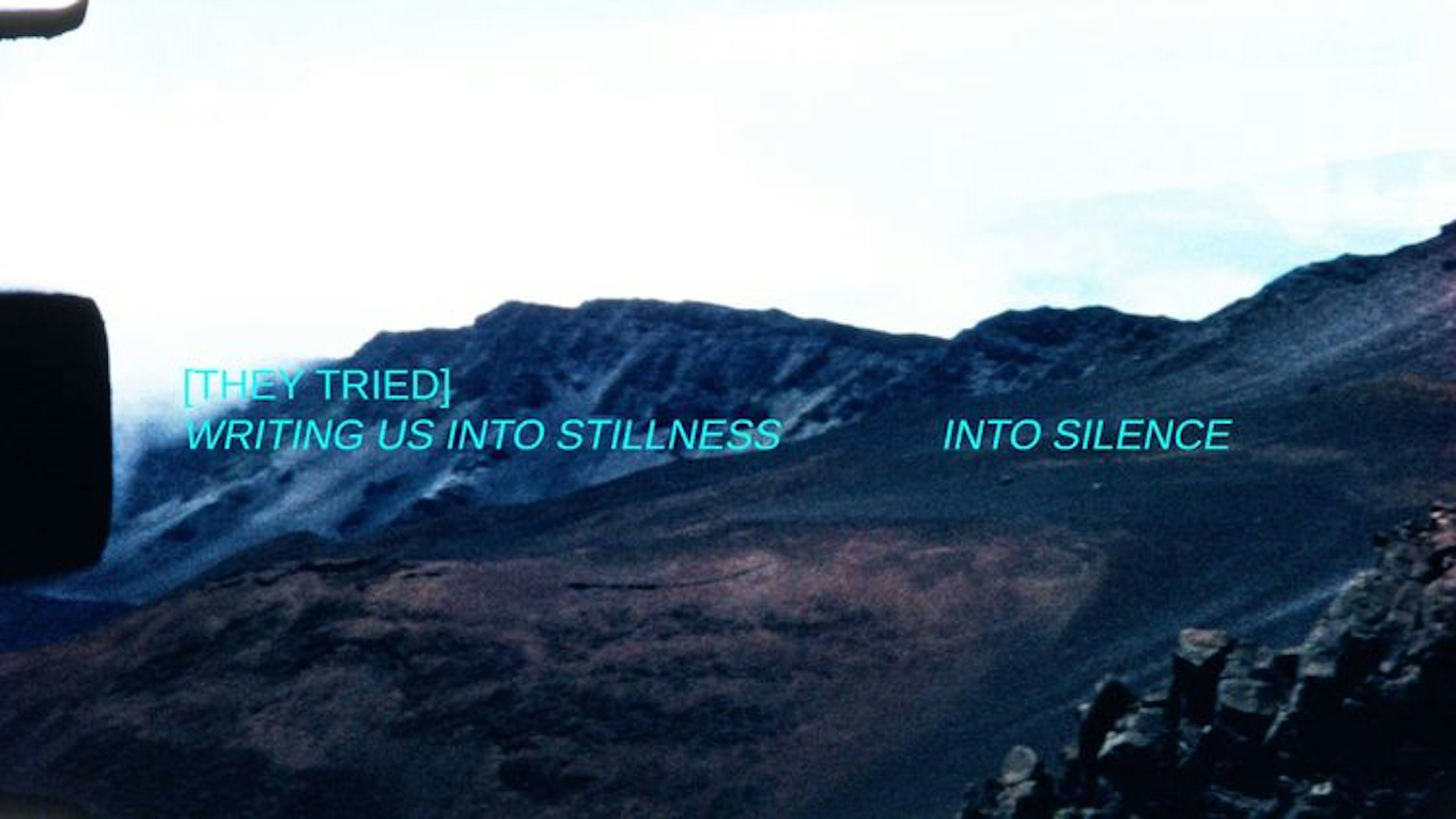 Landscape of mountains with aqua intertitles that read “[they tried] writing us into stillness into silence.” There is a black rectangle on the edge of the frame indicating a perforation from the film scan.
