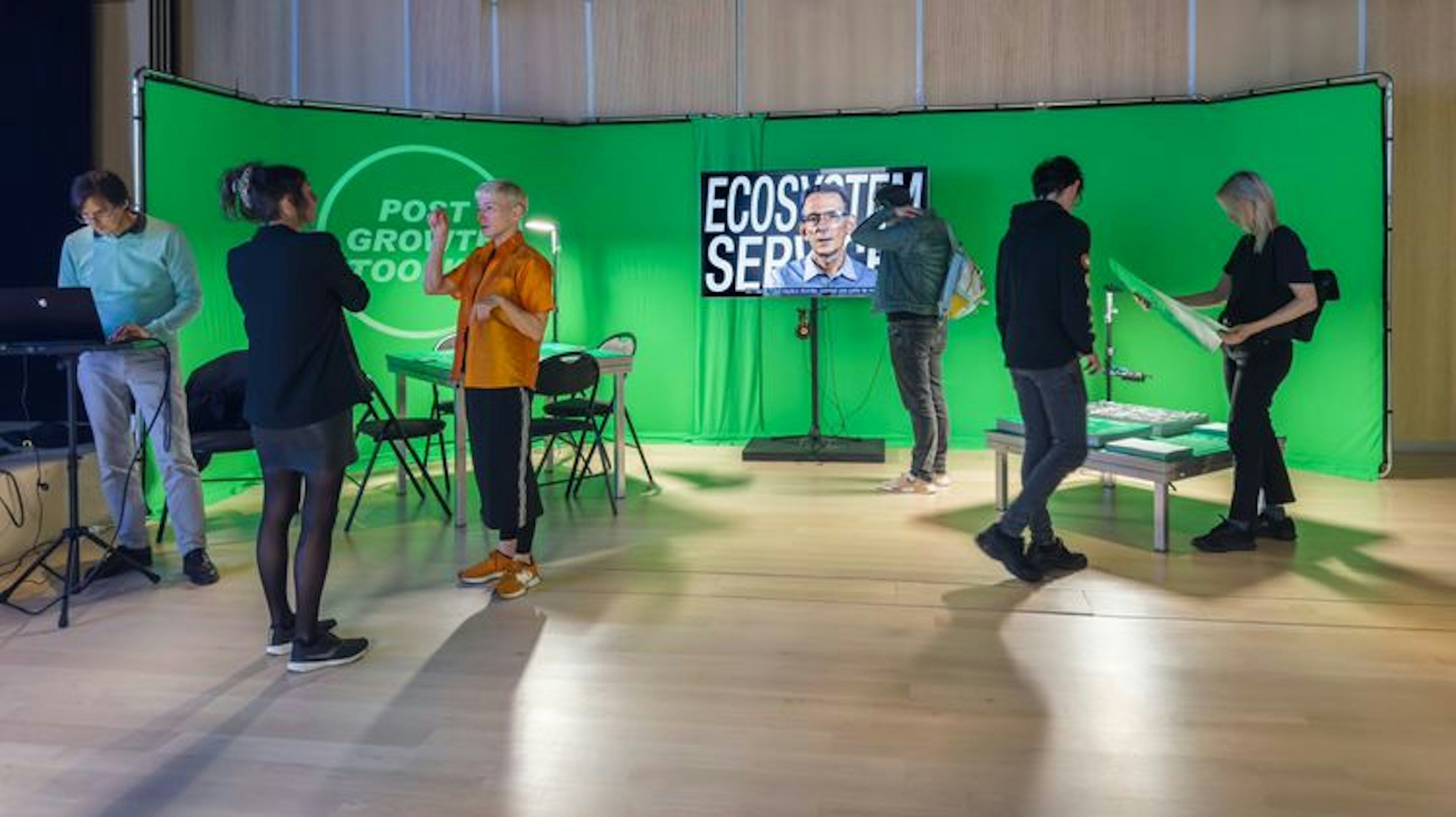 Group of people standing in front of a green screen participating in various activities; conversing, looking at a tv screen, laptop and sheets of paper. 