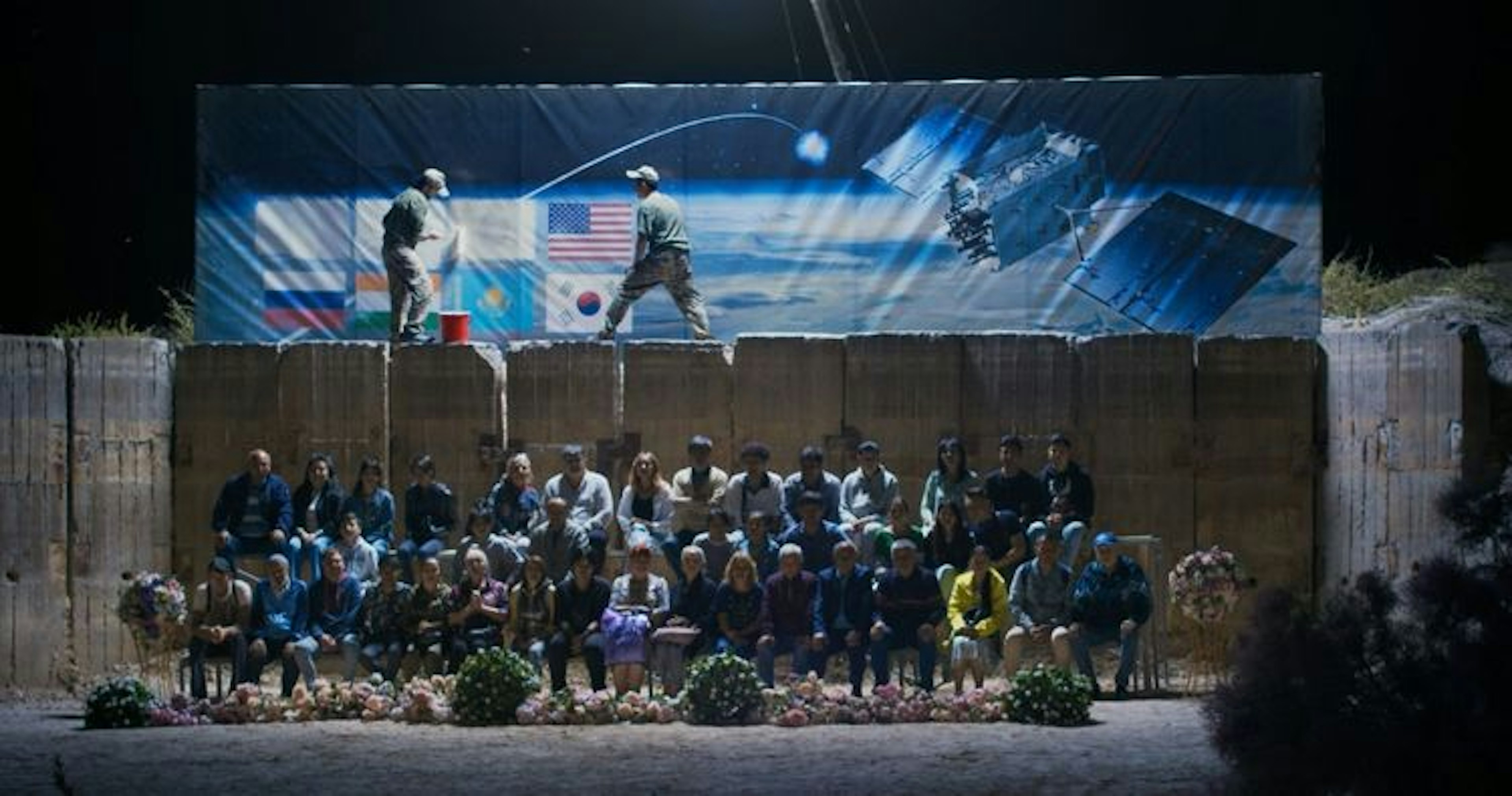A group of people sitting on bleachers viewing a rocket launch at night, while men stand on a wall above the group painting over the flags of countries. 