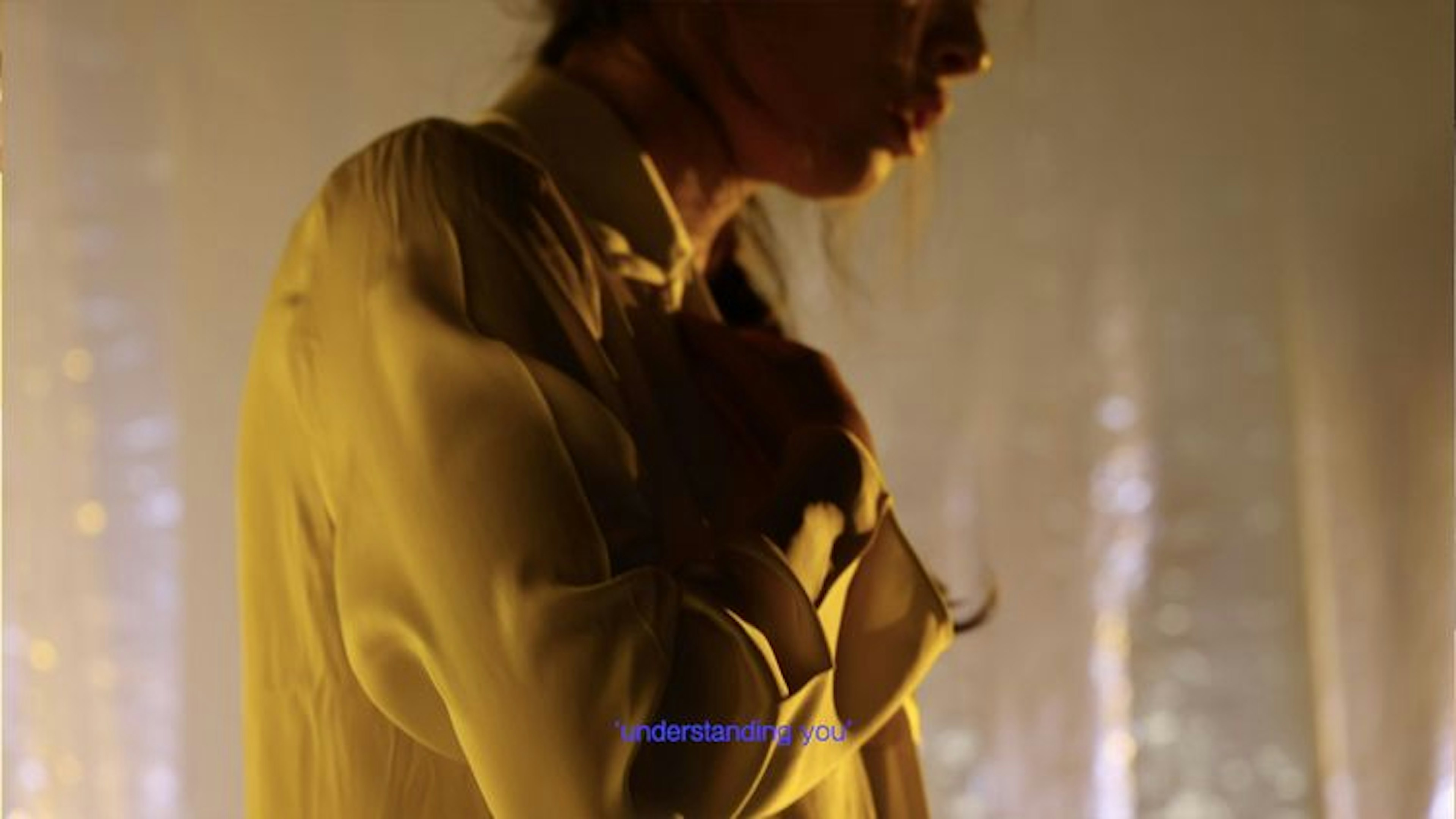 Close-up of a person wearing a yellow chemise, their face slightly out of frame. They have their arms crossed over their chest. There are purple subtitles that read “understanding you.”